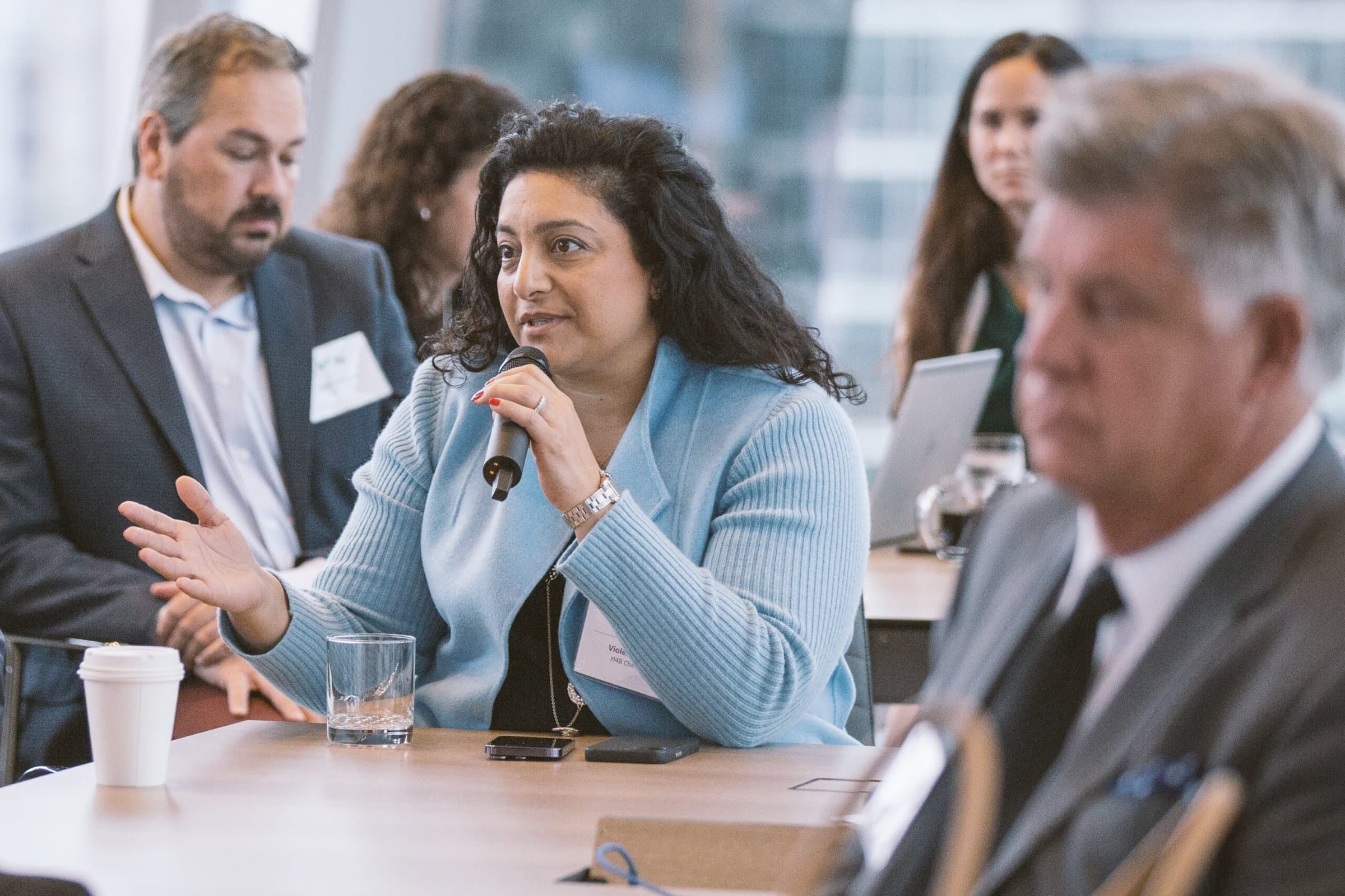 A woman in a blue sweater speaks into a microphone during a meeting or conference. She is seated at a table with a cup of coffee and a smartphone in front of her. Other attendees are seated nearby, listening attentively.