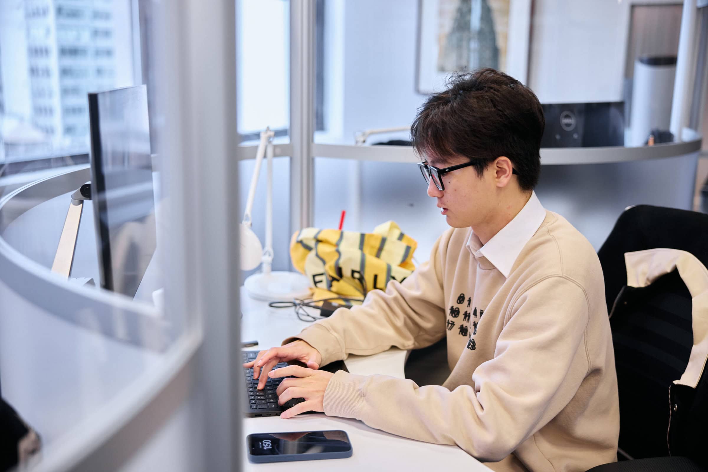 Photo 147 A man typing on a keyboard at a desk in an office, with a yellow bag, lamp, and smartphone on the desk, surrounded by glass partitions.