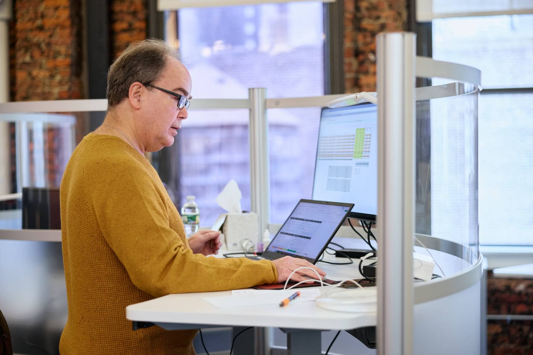 4As_AIHub(1) A man in glasses works at a standing desk in a modern, bright office. He's focused on data displayed on a monitor and laptop in a calm atmosphere.