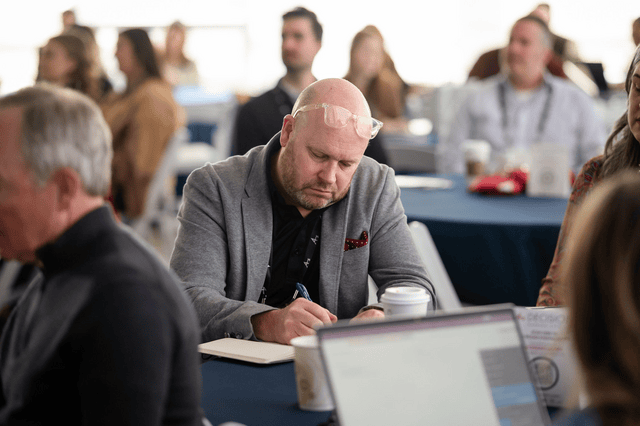 A bald man in a gray blazer writes notes during a conference. He is surrounded by people seated at tables. The atmosphere is focused and professional.
